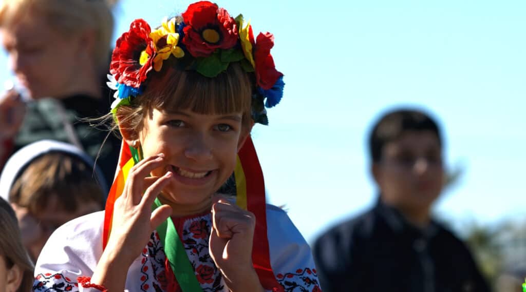 A girl wearing a headband with two ribbons and flowers made of varying colors is awkwardly smiling.